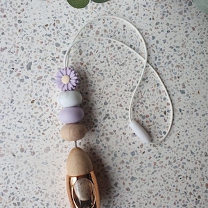 May include: A car air freshener with a gold-coloured, teardrop-shaped base and a wooden top. The freshener has a string of decorative beads in shades of purple, white, and wood tones, topped with a purple flower. A white cord is attached.