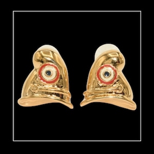 Könnte beinhalten: Ein Paar goldfarbene Ohrringe, jeweils in Form eines stilisierten Helms. Die Ohrringe haben ein rot-weißes kreisförmiges Design mit einem dunkelblauen Zentrum. Der Hintergrund ist schwarz.