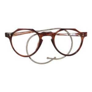May include: Tortoiseshell-patterned eyeglasses with a rounded frame shape and a distinct angular top edge. The glasses feature a thin, silver-toned metal cable or chain that loops behind the frames, connecting to the temples with small metal rivets.