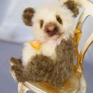 May include: A plush teddy bear with cream-colored fur on its head and chest, and brown fur on its ears, arms, and legs. The bear has dark eyes, a brown nose, and a small orange accent on its chest, sitting on a distressed white wooden chair.