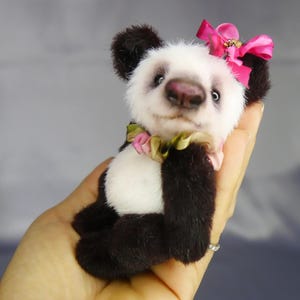 Pode incluir: Um pequeno urso panda de peluche com pelo preto e branco. O panda apresenta um laço de fita cor-de-rosa na cabeça e um colarinho com folhos multicolorido à volta do pescoço. Tem olhos escuros e um nariz escuro, com um aspeto suave.