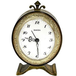 Può includere: Un orologio da tavolo ovale Swiza con quadrante bianco, numeri neri e lancette nere decorate. L'orologio presenta una cornice in metallo dorato con intricati motivi a foglia in rilievo, un fregio decorativo superiore e una base ampia e solida.