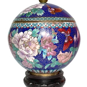 May include: A decorative, egg-shaped cloisonné jar with a lid, featuring a dark blue background and floral and butterfly designs in shades of pink, white, and green. The jar sits on a dark wooden stand and has a brass finial.