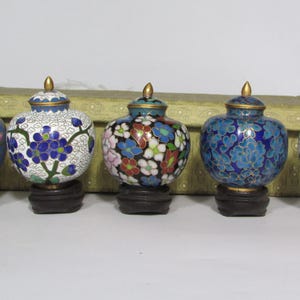 May include: Five decorative cloisonné vases with floral designs in various colors, including blue, white, red, and pink. Each vase has a gold-colored lid and sits on a dark wooden base. The vases are arranged in a row.