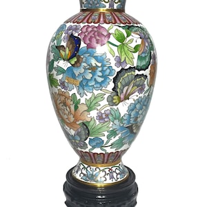 Pode incluir: Um vaso branco de cloisonné com um design floral e de borboletas. O vaso tem uma borda e uma base douradas e assenta sobre um suporte de madeira preto.