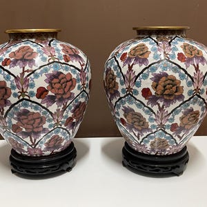 May include: Two matching decorative vases with a white background, featuring a floral pattern in shades of red, purple, and blue. The vases have a gold rim and sit on black wooden stands. The overall design is ornate and detailed.