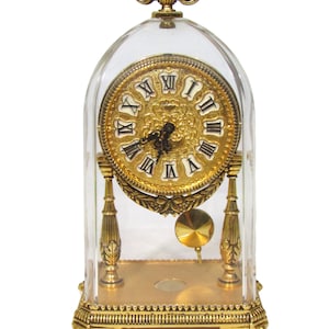 Può includere: Un orologio da camino dorato con numeri romani sul quadrante, un pendolo e una cupola di vetro trasparente. L'orologio è su uno sfondo bianco.