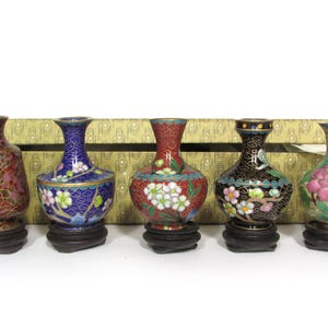 Pode incluir: Cinco vasos decorativos cloisonné com desenhos florais em várias cores, incluindo vermelho, azul e preto. Cada vaso é montado em uma pequena base redonda de madeira escura. Os vasos são exibidos em frente a uma caixa estampada. Altura aproximada: 10 cm.