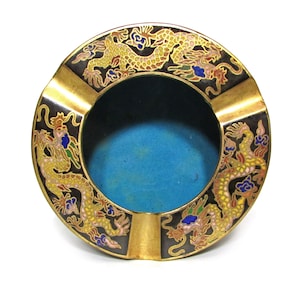 Pode incluir: Um cinzeiro redondo com borda dourada, fundo preto e quatro dragões em esmalte dourado, azul, vermelho e amarelo. Os dragões estão voltados para dentro, em direção ao centro do cinzeiro.