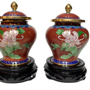 Pode incluir: Dois vasos decorativos cloisonné com tampas. Os vasos são vermelhos com desenhos florais em rosa, verde e branco. Têm detalhes dourados e assentam em bases de madeira preta. As tampas são encimadas por pináculos dourados.