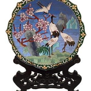 Pode incluir: Um prato decorativo redondo de cloisonné com uma paisagem serena com guindastes, flores de cerejeira e montanhas. O prato está sobre um suporte de madeira escura. A borda do prato é verde com detalhes florais.