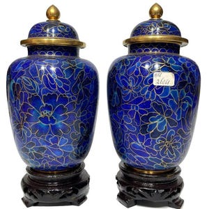 Op de afbeelding: Twee blauwe decoratieve vazen met deksels, versierd met een bloemmotief in blauw en goud. Ze staan op donkere houten voetstukken. De deksels hebben gouden knoppen en randen. De vazen zijn ongeveer 30 cm hoog.