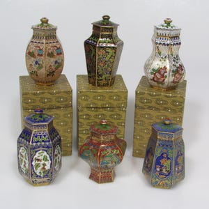 May include: Six small, octagonal, cloisonné enamel boxes with lids. The boxes are decorated with floral and geometric patterns in shades of blue, green, red, gold, and white. The boxes are displayed on small, square, gold boxes with a cloud pattern.