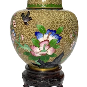 Può includere: Un vaso decorativo cloisonné vintage con coperchio, posto su un supporto in legno scuro. Il vaso presenta uno sfondo beige con un motivo a nuvole ed è decorato con un motivo floreale rosa, blu e verde e un piccolo uccello. Il coperchio ha un bordo verde.