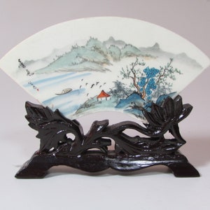 Puede incluir: Un abanico de porcelana blanca con una escena de paisaje pintada de montañas, agua y árboles. El abanico se muestra en un soporte de madera negra con tallas intrincadas.
