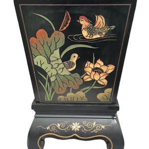 Puede incluir: Una maceta lacada en negro con parte superior acampanada y base de pedestal. El panel frontal presenta una escena pintada a mano de dos pájaros, una gran hoja de loto verde y una flor de loto naranja. La base está decorada con motivos florales dorados.