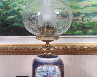 May include: A vintage porcelain lamp base with a blue and white floral design and gold accents. The lamp has a clear glass shade with etched details and a frosted glass chimney.