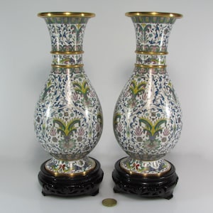 May include: A pair of cloisonné vases with a white background and intricate floral patterns in shades of blue, green, yellow, and pink. The vases have a gold rim and are sitting on black wooden stands.