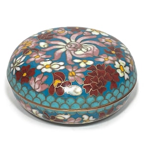 May include: A round, decorative cloisonné box with a floral design. The lid features a turquoise background with red, pink, and white flowers. The box has a gold-colored metal rim and a turquoise base with a scale pattern.