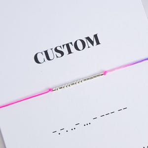 Può includere: Un biglietto bianco con la parola "CUSTOM" stampata in nero. Un braccialetto di corda rosa e blu con un ciondolo d'argento è sul biglietto. Il ciondolo ha una serie di trattini che rappresentano un messaggio in codice Morse. Il biglietto dice anche "925 SILVER MORSE CODE BRACELET" e "@florinejewelry".