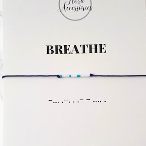 Puede incluir: Un brazalete de cuentas azul y blanco con la palabra "BREATHE" y su representación en código Morse en una tarjeta blanca. La tarjeta también incluye el texto "Pulsera de código Morse" y una dirección de sitio web.