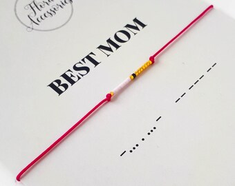 Könnte beinhalten: Ein rotes Armband aus Schnur mit weißen, gelben und schwarzen Perlen, die "BEST MOM" in Morsecode buchstabieren. Das Armband befindet sich auf einer weißen Karte mit dem Text "MORSE CODE BRACELET" und einer Website-Adresse.