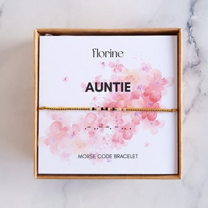 Op de afbeelding: Een gouden armband met zwarte kralen die "AUNTIE" in morsecode spellen. De armband zit in een witte doos met een roze bloemenontwerp en de woorden "florine" en "MORSE CODE ARMBAND".