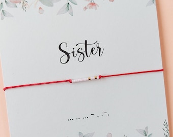 May include: Red string bracelet with white and gold beads. The bracelet is on a card with a floral design and the word "Sister" written in cursive. The card also includes the Morse code for "Sister" and the website address for the shop.
