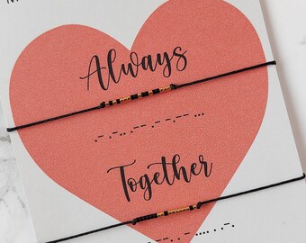 May include: A pair of black string bracelets with gold beads spelling out "Always" and "Together" in Morse code. The bracelets are displayed on a white card with a pink heart and the text "Morse Code Couple Bracelet".