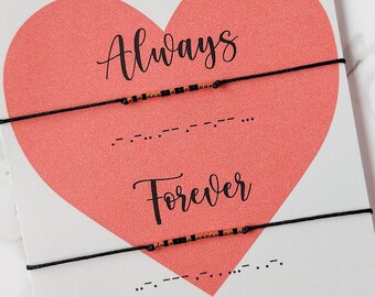 May include: A pair of black string bracelets with small beads. The bracelets are displayed on a white card with a pink heart and the words "Morse Code Couple Bracelet", "Always", and "Forever" in black text. The words "Always" and "Forever" are written in Morse code on the card.