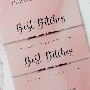 Op de afbeelding: Twee vriendschapsarmbanden met de tekst "Best Bitches" in zwarte cursieve letters op een roze achtergrond. De armbanden zijn gemaakt van zwarte en roze kralen en worden getoond op een kaart met de tekst "Morse Code Armband" en het websiteadres "www.floraaccessorieshop.etsy.com".
