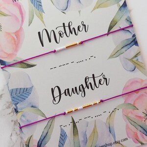 May include: Two purple string bracelets with gold beads spelling out "Mother" and "Daughter" in Morse code on a white card with a floral design. The card also includes the text "Morse Code Bracelet" and a website address.