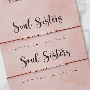 Könnte beinhalten: Zwei braune Lederarmbänder mit weißen und goldenen Perlen, die "Soul Sisters" in Morsecode auf einer rosa Karte mit dem Text "Morsecode Armband" buchstabieren.