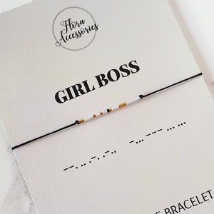 Peut inclure: Un bracelet en cordon noir avec des perles blanches et dorées qui épellent "GIRL BOSS" en code Morse. Le bracelet est sur une carte blanche avec le texte "GIRL BOSS" et "MORSE CODE BRACELET" en noir.