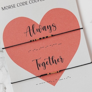 May include: A pair of black string bracelets with gold beads spelling out "Always" and "Together" in Morse code. The bracelets are displayed on a white card with a pink heart and the text "Morse Code Couple Bracelet".