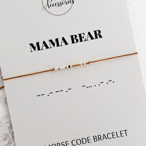 Peut inclure: Un bracelet en cordon marron avec des perles blanches et dorées qui épellent "MAMA BEAR" en code Morse. Le bracelet est présenté sur une carte blanche avec le texte "MAMA BEAR" et "BRACELET CODE MORSE" ainsi que l'adresse du site Web "www.floraaccessorieshop.etsy.com".