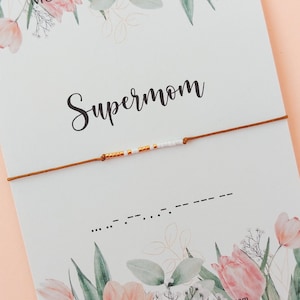 Op de afbeelding: Een witte kaart met een bloemenpatroon en de tekst "Morse Code Bracelet" en "Supermom". Een bruin koordarmband met witte en gouden kralen ligt op de kaart. De morsecode voor "Supermom" staat onder het armband.