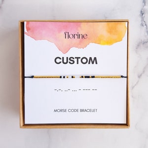 Può includere: Un biglietto bianco con uno sfondo ad acquerello in tonalità rosa e arancio. Il biglietto dice "florine" e "CUSTOM" in testo nero. Sotto la parola "CUSTOM" c'è un braccialetto di perline dorate e nere con uno stringa nera. Il testo "BRACCIALE CODICE MORSE" è stampato sotto il braccialetto.