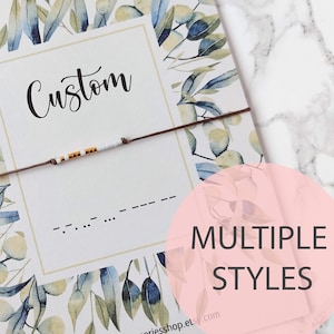 Op de afbeelding: Een witte kaart met een aquarel bloemenrand en de tekst "Morse Code Bracelet" bovenaan. De kaart heeft ook de tekst "Custom" in een handschrift en een regel morsecode. De kaart ligt op een marmeren oppervlak. De tekst "MULTIPLE STYLES" staat in een roze cirkel.