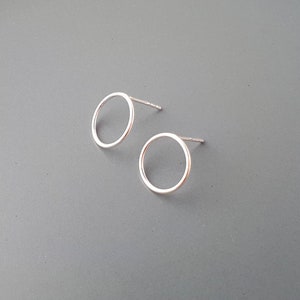 Op de afbeelding: Twee zilverkleurige oorstekers met een open cirkel. De oorbellen hebben een minimalistisch ontwerp. Ze zijn klein en geplaatst tegen een grijze achtergrond, wat hun eenvoudige, moderne esthetiek benadrukt. Een klassiek sieraad.