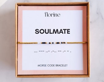 Puede incluir: Una caja blanca con un interior rosa y un brazalete de cuerda dorada en su interior. La caja tiene las palabras "florine" y "SOULMATE" impresas en ella. El brazalete tiene cuentas negras y blancas y las palabras "PULSERA DE CÓDIGO MORSE" están impresas debajo del brazalete.