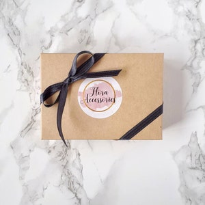 May include: A brown cardboard box with a black ribbon tied around it. The box has a white circle sticker with the text "Flora Accessories" in pink lettering.
