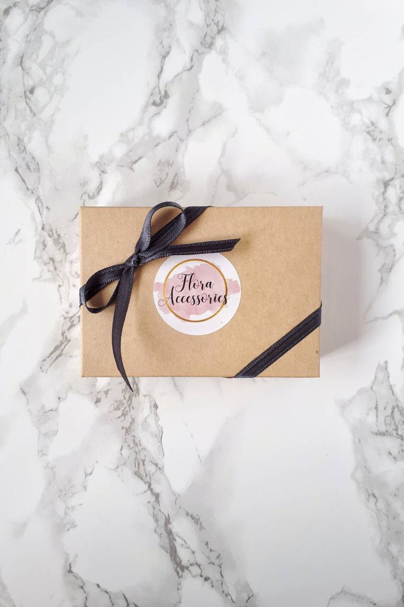 May include: A brown cardboard box with a black ribbon tied around it. The box has a white circle sticker with the text "Flora Accessories" in pink lettering.