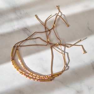 Puede incluir: Tres pulseras superpuestas con cuentas rosas y doradas y lazos de cuerda marrón.