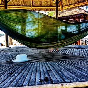 以下が含まれることがあります： 木製のデッキに、2本の木製の支柱の間に緑色のハンモックが吊り下げられています。ハンモックの前に、白い帽子がデッキの上に置かれています。