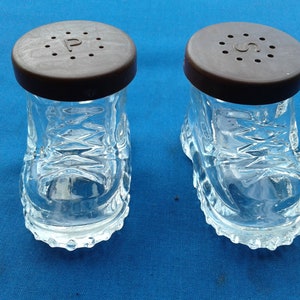 May include: A pair of glass salt and pepper shakers shaped like boots. The shakers have brown plastic lids with holes for dispensing salt and pepper. The lids have the letters "P" and "S" printed on them.