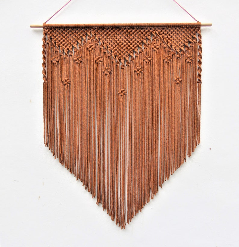 May include: A brown macrame wall hanging. The top section features a woven design, with vertical strands cascading downwards. The bottom edge is cut at an angle, creating a pointed shape. The wall hanging is suspended from a wooden dowel.