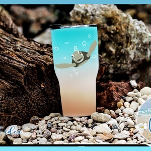 May include: A turquoise and peach ombre tumbler with a graphic of a sea turtle swimming in the ocean. The tumbler is sitting on a bed of small white rocks.