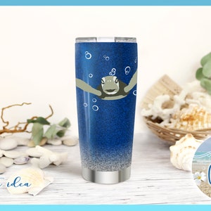 May include: A blue and silver stainless steel tumbler with a sea turtle design and bubbles. The tumbler is sitting on a white wooden surface with seashells and a sign that says "Sea Craze".