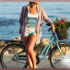 May include: A woman in a white tank top with the text "sand, saltwater, sun" and a blue and white graphic design stands next to a blue bicycle with a basket of flowers. The bicycle is parked on a wooden boardwalk near the ocean.